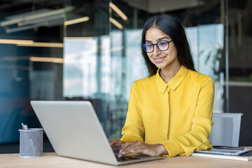 Smiling and cheerful Indian businesswoman wearing glasses and a yellow shirt working in the office on a laptop