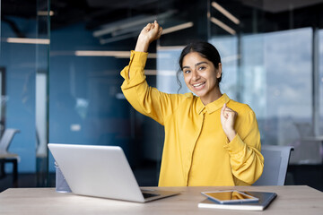 Portrait of a young Indian woman sitting at a desk in the office, rejoicing and dancing, looking at the camera with a smile