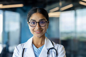 Close-up portrait of a young Indian female doctor. scientist in a white coat and glasses. standing indoors, looking and smiling at the camera