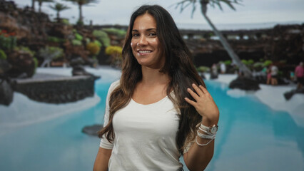 Woman smiling and touching her hair at a turquoise pool with palm trees and lava rock terrace in an outdoor resort setting; vacation contentment.