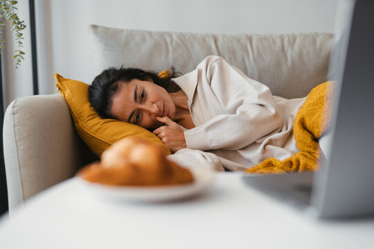 Thoughtful young woman lying on a cozy sofa at home, resting her head on a yellow pillow while looking ahead quietly, wrapped in a warm blanket with breakfast on the table.