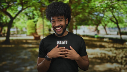 Man in black uniform smiling with hands on chest in green outdoor park with sunlight filtering through trees creating a peaceful background setting.