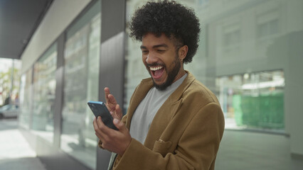 African american man on city street using smartphone with joyful expressions, showcasing urban outdoor lifestyle and technology engagement.