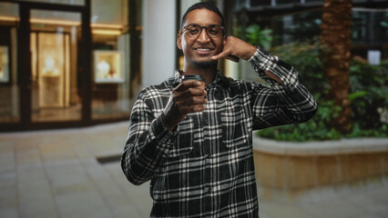 Young man wearing plaid shirt and glasses holding a takeaway coffee cup and making a phone gesture...