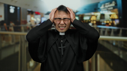 Obraz premium Priest wearing black robe and glasses in a mall expresses stress while holding head with both hands in front of blurred indoor shopping center background
