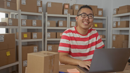 Man in a red striped shirt typing on laptop with parcel at a packing desk with rows of boxes and...