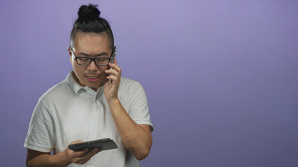 Young man wearing glasses and white polo talking on phone while holding a tablet in purple studio; frustration.