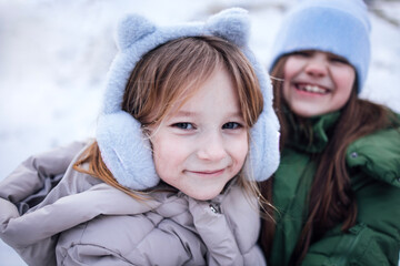 Fototapeta premium Two young girls, one wearing gray ear muffs and the other in a blue hat, are smiling joyfully in a snowy outdoor setting, capturing the essence of winter fun and friendship