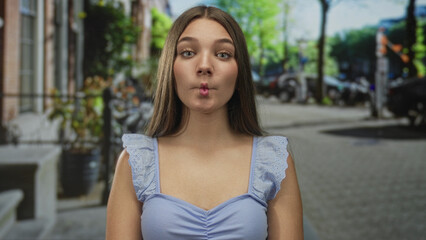 Teenager girl puckers lips and tilts head slightly in a light blue top with exposed shoulders on a sunny urban street scene; playfulness.