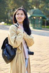 Naklejka premium Woman with camera and backpack stands in a sunny park, wearing a beige trench coat and soft scarf, ready for outdoor photography, exploration, and casual storytelling; natural light highlights her