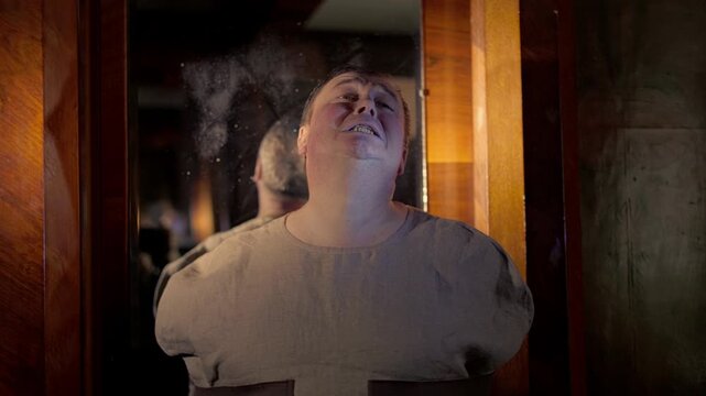 Mentally ill person.A dim room. A man stares blankly into his own reflection. He is wearing a white straitjacket with long, tied sleeves. The man is crying delirium tremens.