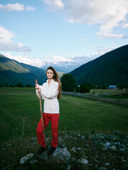 Naklejka premium Mountain field outdoor scene with a woman in red pants holding a staff, valley views, clear sky and rustic fence, capturing nature and exploration in wide rural landscape