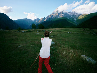 Naklejka premium Back view of a hiker in a white top and red pants standing in a wide meadow with rocks, pointing toward distant mountains under a bright blue sky and clouds