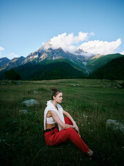 Naklejka premium Woman sits in a green meadow at the foot of towering mountains, wearing red pants and a white top, gazing into the distance under a blue sky with fluffy clouds during outdoor travel