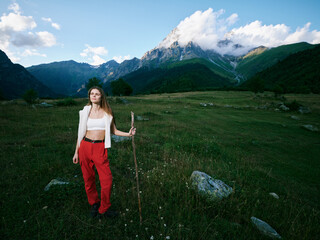 Naklejka premium Woman standing in a mountain meadow with a walking stick, wearing red pants and a white top, expansive landscape with distant peaks and open outdoors