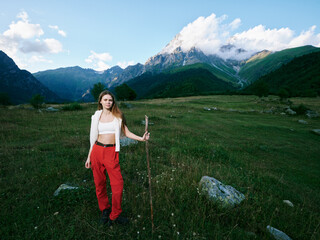 Naklejka premium Woman with a wooden staff stands in a green meadow beneath towering mountains, wearing a white jacket and red pants, capturing a tranquil alpine hiking scene amid wildflowers