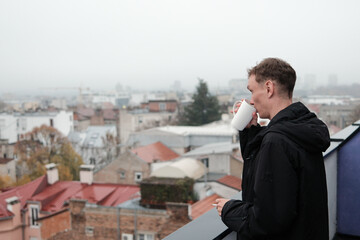 A man drinks coffee on a terrace while holding a cigarette and looking over the foggy city. The scene reflects a quiet morning concept.