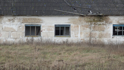 Abandoned building with broken windows surrounded by overgrown grass on a cloudy day in a rural area