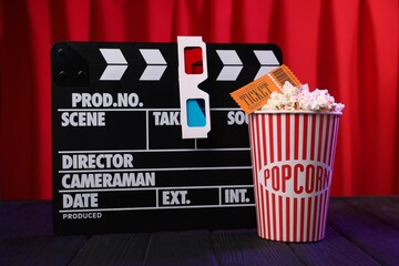 Tasty popcorn in paper cup, movie ticket and clapboard on purple wooden table against red curtains