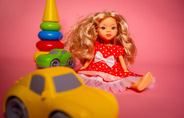 A doll with curly blonde hair wears a red dress with white dots. Nearby, bright toy cars and a stacking toy are arranged on a pink surface in a fun space.