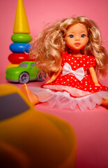 A doll with curly blonde hair wears a red dress with white dots. Nearby, bright toy cars and a stacking toy are arranged on a pink surface in a fun space.