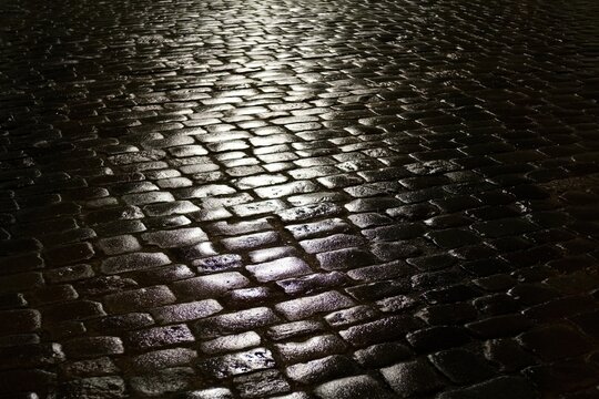 Wet cobblestone pavement reflecting streetlight at night, creating a moody urban texture and atmospheric glow.
