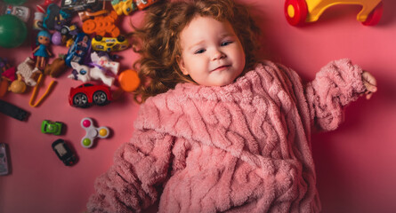 A young child lies on a pink floor surrounded by colorful toys. The child smiles widely while wearing a cozy sweater. There are many toy cars and figures scattered around.