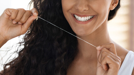 A woman with curly hair is smiling as she flosses her teeth. The warm lighting creates a friendly atmosphere, highlighting her focus on dental care.