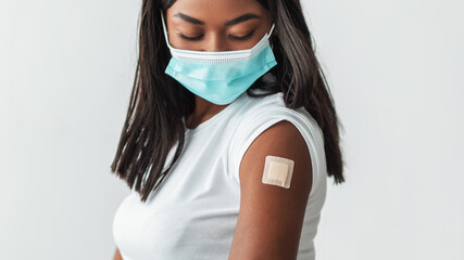 Vaccination For Protection During Global Covid-19 Pandemic. Portrait of black millennial female patient in face mask showing arm with sticking plaster after injection posing on white studio background