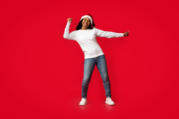 A joyful young woman in a Santa hat dances playfully in front of a vibrant red backdrop. She wears a casual white shirt and jeans, celebrating the festive spirit.