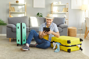 Happy man traveler choosing tour for holiday trip sitting at home with packed suitcases and booking tickets online via tablet. Young tourist getting ready for voyage. Vacation and travelling concept.