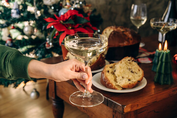 A woman's hand holds an elegant glass of sparkling wine against the background of a festive Christmas table with traditional Italian panettone beautifully decorated Christmas tree in the background