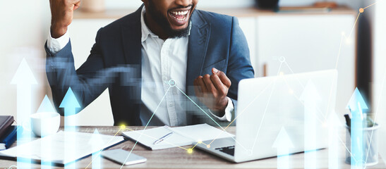 A happy black businessman is celebrating his success at work in a modern office. He expresses joy while looking at his laptop screen, surrounded by business materials and graphics showing growth.