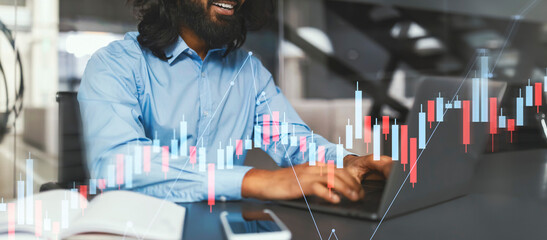 A smiling man with long hair and glasses is focused on his laptop at his office desk. Charts and financial graphics are visible on the screen, indicating he is working on data analysis.