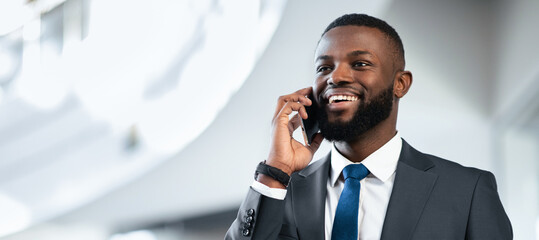 A man is using a mobile phone while smiling in an office space. He wears a suit and looks engaged in the conversation. Light enters through windows creating an open atmosphere.