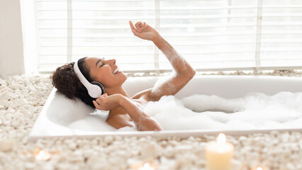 A woman enjoys a relaxing bubble bath in a stylish tub surrounded by stones and candles. She is smiling and dancing to music with headphones on, creating a tranquil atmosphere.