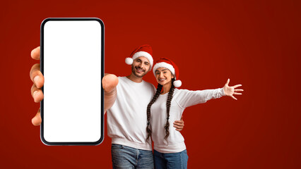 A joyful couple is standing together in front of a bright red background. They both wear Santa hats and show excitement while holding a smartphone with a blank screen.