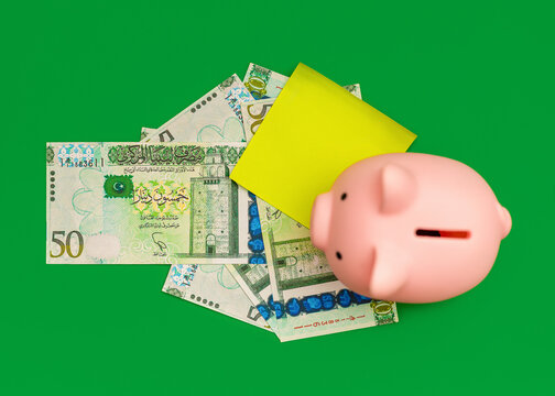 A pink piggy bank, a neon yellow sticky note, and several green banknotes of 50 Libyan Dinar,  are arranged on a bright green background.