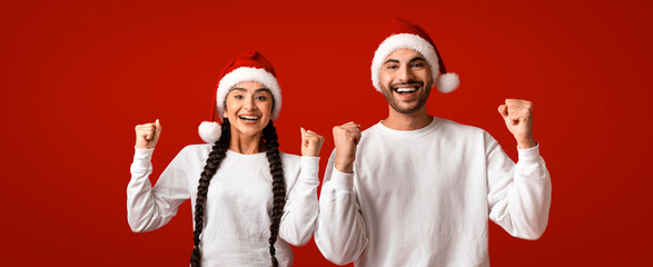 Two friends, one with braids and the other with a beard, joyfully celebrate the holiday season in matching white shirts and Santa hats against a bright red background.