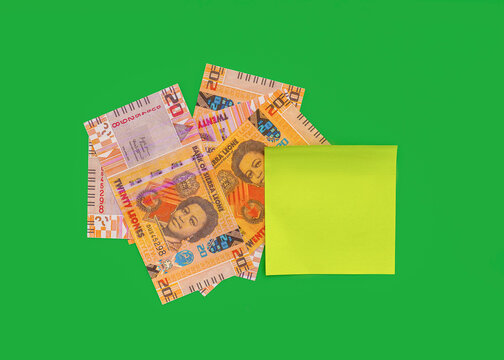 A cluster of 20 Sierra Leonean Leones banknotes, orange and red, scattered and partially covered by a blank, square yellow-green sticky note against a bright green background.