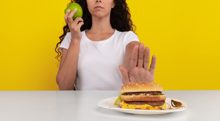 A woman with curly hair holds an apple in one hand while pushing away a hamburger on a plate with the other hand. The vibrant yellow background highlights her choice for healthier eating.