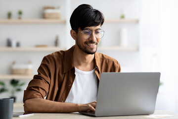 A young man sits at a wooden table in his home office, focused on his laptop. Soft sunlight fills the space, creating a warm and inviting atmosphere.