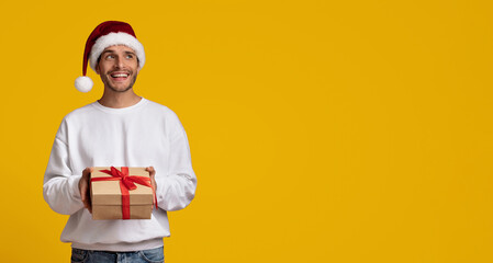 A smiling man wearing a Santa hat stands holding a wrapped gift box with a red ribbon in front of a vibrant yellow background. His cheerful expression suggests happiness and excitement.