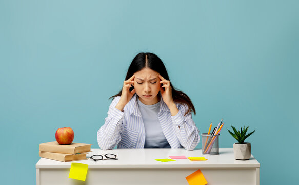 A young woman sits at her study desk with a focused expression, rubbing her temples as she deals with stress. Books, stationery, and an apple surround her.