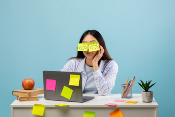 A woman looks tired as she covers her eyes with sticky notes featuring drawn eyes. She sits at a...