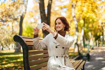 A woman with red hair is smiling as she takes pictures with her smartphone in a scenic park. The trees surrounding her are vibrant with autumn colors.