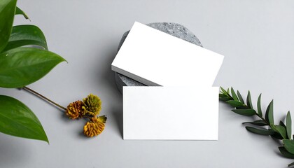 Two blank white business cards mockup with natural green leaves and dried flowers on a light grey background, perfect for branding.