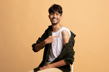 A young man sits in a bright studio, smiling widely while pointing at the bandage on his arm. He has short hair and wears glasses, with a casual outfit that includes a tank top and open shirt.