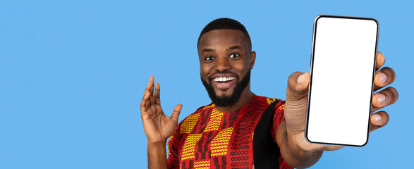 A man wearing colorful traditional clothing stands with a cheerful expression, holding up a blank smartphone. He appears to be promoting a product or sharing news. The backdrop is a solid blue color.