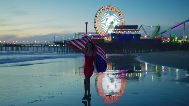 Girl walks along Santa Monica beach at sunset, USA flag in hand